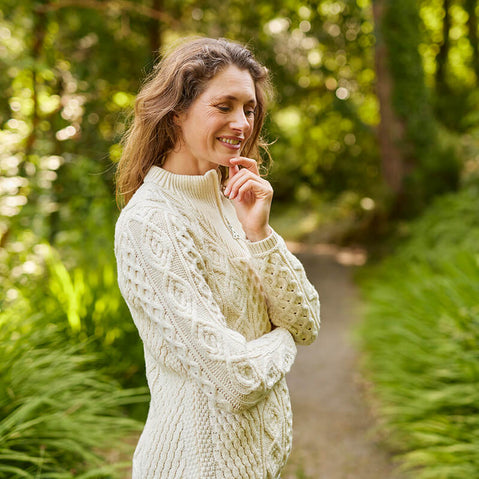 An irish woman in a cream Aran sweater stands on a pathway surrounded by greenery. She smiles softly while resting her chin on her hand, appearing thoughtful and serene.
