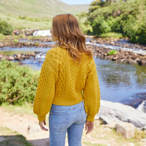A woman stands with her back to the camera, wearing a yellow knitted Aran sweater and light blue jeans. She gazes over a serene river surrounded by lush greenery and rocky terrain. The sunlight illuminates the scene, enhancing the natural landscape.