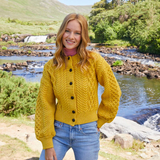 A woman stands by a river, smiling and wearing a yellow knitted Aran cardigan with buttons. She pairs it with blue jeans, and the backdrop features greenery and a waterfall.