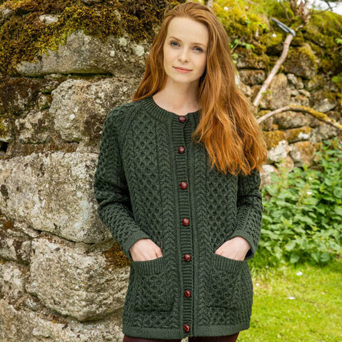 A woman with long, wavy red hair stands in front of a stone wall covered in moss. She wears a dark green knit cardigan with a textured honeycomb stitch pattern and button closure, featuring pockets on the front. The background includes lush greenery.