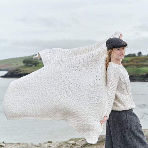 Irish home décor- Traditional Aran Honeycomb Wool Throw. A person stands on a rocky coastline, wearing a light-colored knitted sweater and dark trousers. They hold a large, cream Aran knitted blanket open behind them, creating a flowing effect against a backdrop of rolling hills and water. The sky is cloudy and the scene suggests an outdoor environment with natural elements.