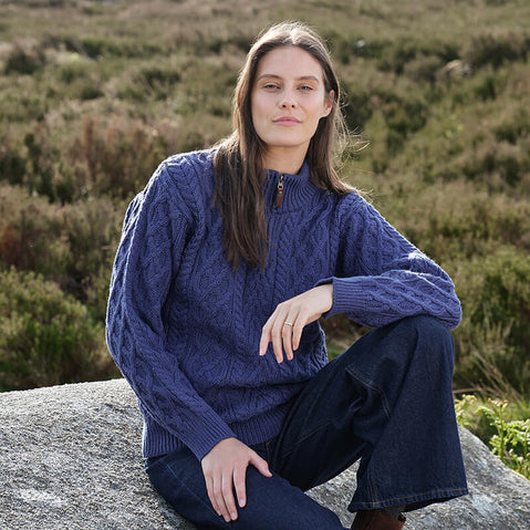Person wearing a blue Women's Signature Half Zip Irish Wool Sweater, featuring intricate cable knit patterns and a half-zip collar, sitting on a rock in an outdoor setting with greenery in the background