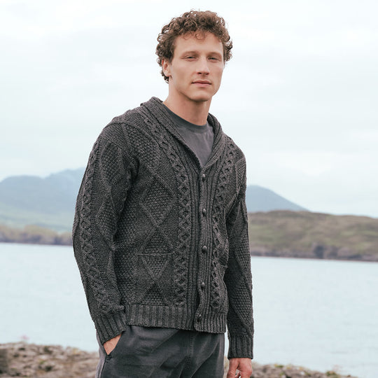 Image featuring the Men's Heritage Aran Shawl Collar Button Cardigan in charcoal, crafted with authentic Irish Aran cable knit patterns, a timeless shawl collar, and classic button-front styling, showcasing traditional Irish craftsmanship against a rugged coastal landscape.
