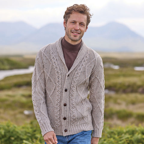 Man wearing an oatmeal Men's Heritage Aran Shawl Collar Button Cardigan in a scenic outdoor setting with mountains and fields.
