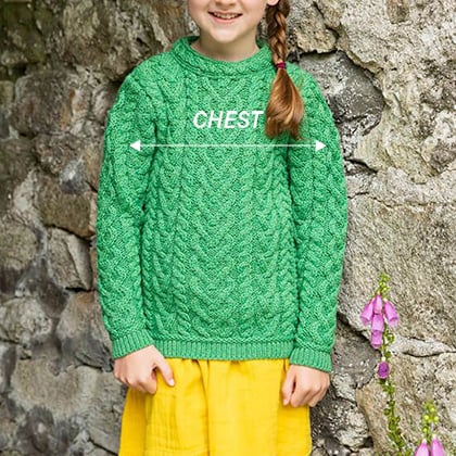 A young girl stands in front of a stone wall, wearing a bright green cable knit sweater and a yellow skirt. The word