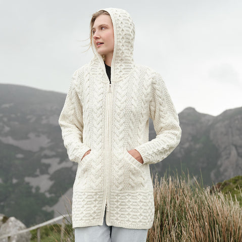 Image featuring the Hooded Aran Zipper Classic Coat in cream, crafted with authentic Irish Aran cable knit patterns, a practical hood, full-zip closure, and spacious front pockets, showcasing timeless Irish craftsmanship in a scenic mountain setting.