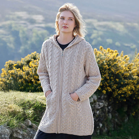 Woman wearing a beige Aran cardigan standing outdoors with greenery and flowers in the background