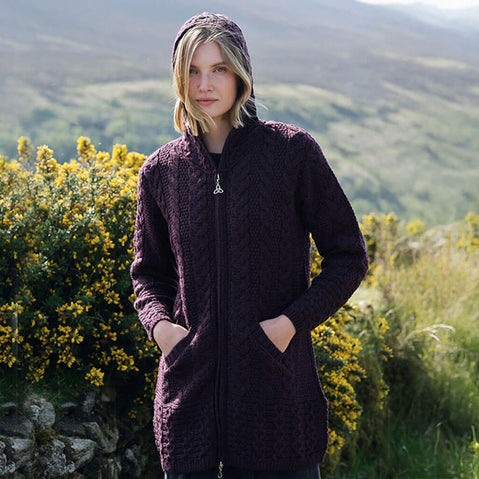 Woman wearing a dark purple Aran zipper cardigan standing in a scenic outdoor setting with green hills and yellow flowers.