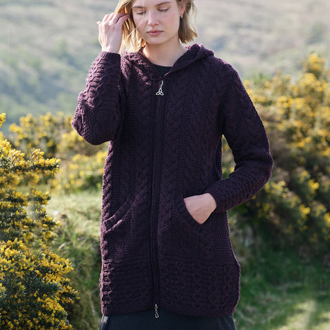Woman wearing a dark purple knitted Aran cardigan with a hood in a natural setting with greenery.