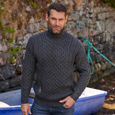 Man wearing a charcoal Men's Signature Fisherman Turtleneck Wool Sweater standing in front of a stone wall.
