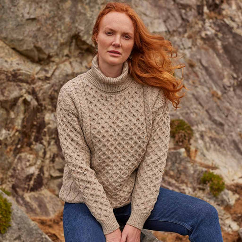 Woman wearing an oatmeal Women's Signature Fisherman Turtleneck Wool Sweater and blue jeans against a rocky background