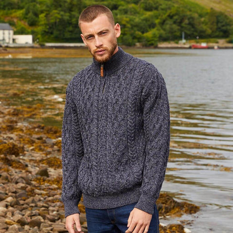 Man wearing a dark blue knitted sweater standing by a body of water with greenery in the background