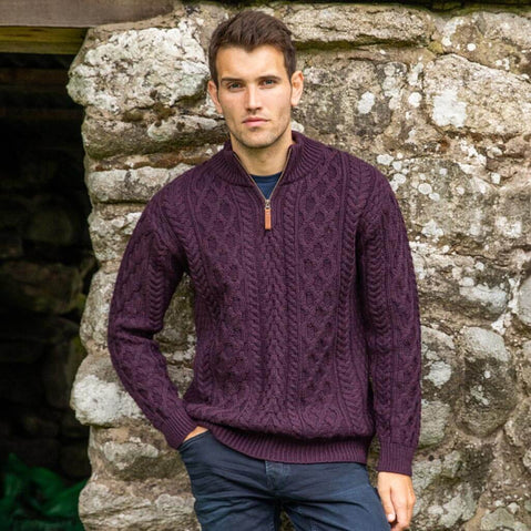 Man wearing a purple cable knit sweater standing against a stone wall.