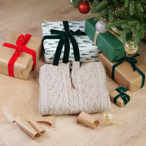 Beige cable knit sweater folded under Christmas tree with wrapped gifts in festive green and gold paper with ribbon bows
