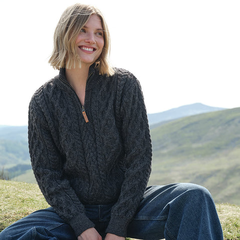 Woman wearing a dark gray cable knit sweater sitting on a grassy hill with mountains in the background