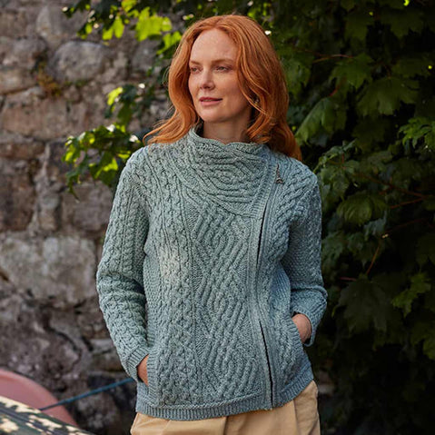 Woman wearing an aqua Women's Signature Aran Wool Zipper Cardigan standing against a stone wall with greenery.