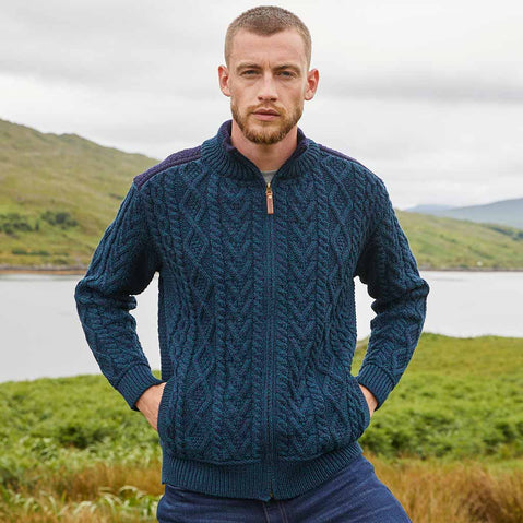 Man wearing a blackwatch blue Men's Fisherman Full-Zip Aran Cardigan standing in a scenic outdoor setting with mountains and water.