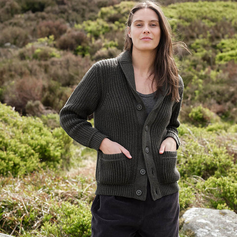 Woman wearing a dark green Aran cardigan in a natural setting with greenery.