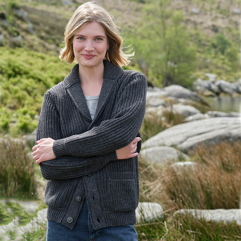 Woman wearing a dark gray Aran cardigan in a natural setting with greenery and rocks.