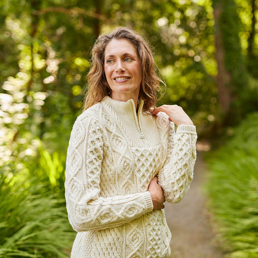 A woman stands on a path surrounded by lush greenery, wearing a cream Aran sweater. She smiles softly while holding her arm, exuding a relaxed and cheerful demeanor in an outdoor setting.