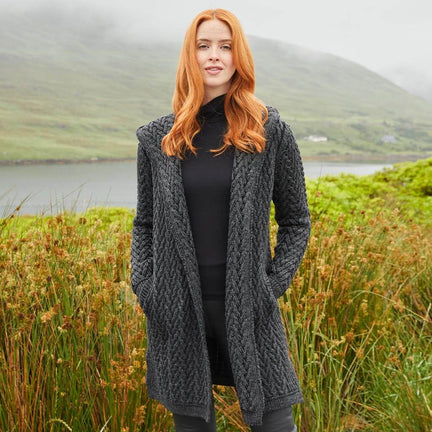 Woman wearing an Irish Aran waterfall cardigan in charcoal gray standing by a lake with lush greenery and misty mountains in the background.