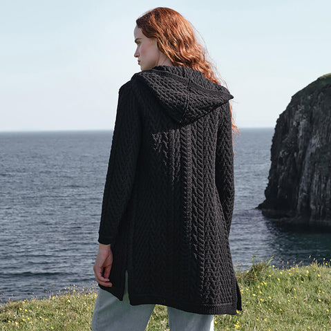 Back view of the Women's Signature Hooded Aran Waterfall Cardigan in Black, showcasing intricate traditional Irish cable knit patterns and a textured hood, styled along the dramatic Irish coastline with rugged sea cliffs and ocean views in the background.