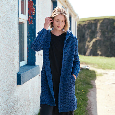 Women's Signature Hooded Aran Waterfall Cardigan in Royal Blue, featuring traditional Irish cable knit stitching and a relaxed open-front design with hood, styled beside a traditional Irish cottage along the rugged coastline.