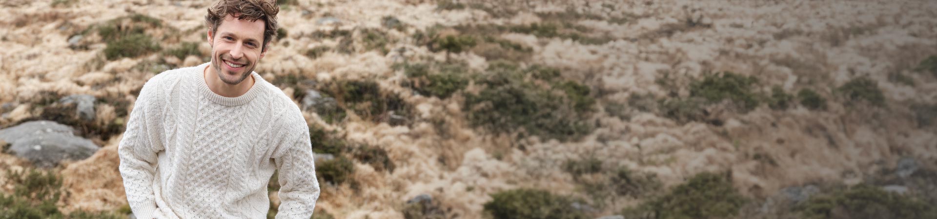 A smiling man in a white knit aran sweater sits on a grassy hillside, with a natural landscape of muted colors and rocks in the background.