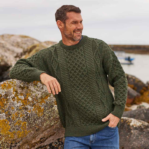 Man wearing an army green Men's Fisherman Lightweight Crew Neck Heritage Sweater standing on a rocky shoreline.