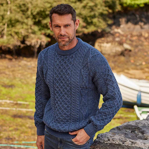Man wearing a blue Men's Fisherman Lightweight Crew Neck Heritage Sweater standing outdoors with trees and water in the background
