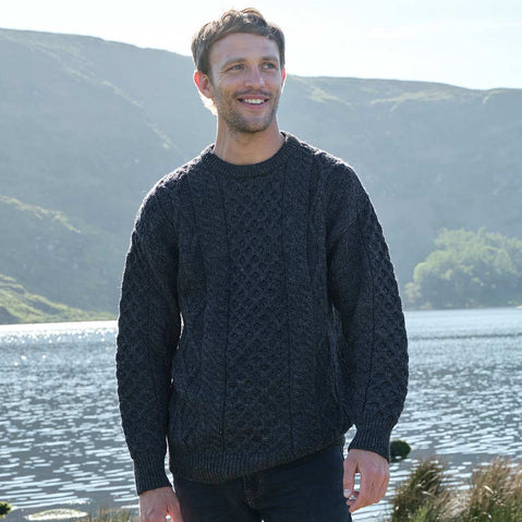 Man wearing a charcoal Men's Fisherman Lightweight Crew Neck Heritage Sweater standing by a lake with mountains in the background