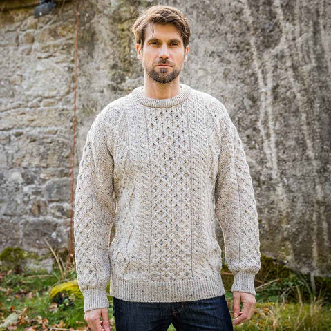 Man wearing an oatmeal Men's Fisherman Lightweight Crew Neck Heritage Sweater standing in front of a stone wall.