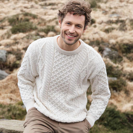 Man wearing a cream Men's Fisherman Lightweight Crew Neck Heritage Sweater sitting outdoors with a natural landscape in the background