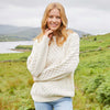 Woman wearing a cream Womens Fisherman Lightweight Crew Neck Heritage Sweater standing in a scenic outdoor setting with a lake and mountains.