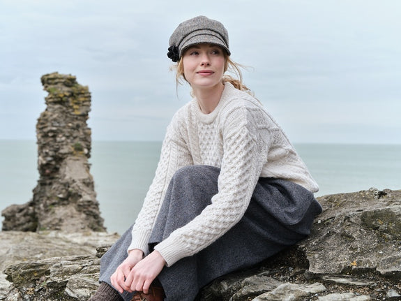 A young woman sits on a rocky shoreline, wearing a white knitted aran sweater and a gray skirt. She has a tweed cap on her head and gazes into the distance, with an ocean and cloudy sky in the background.