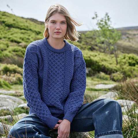 Woman wearing a blue Aran sweater sitting in a natural setting with green hills.