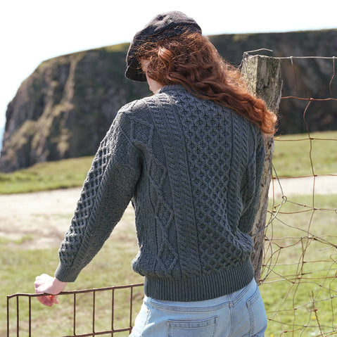 Back view of the Women's Fisherman Lightweight Crew Neck Heritage Sweater in Lichen, highlighting intricate traditional Irish cable knit patterns and authentic heritage craftsmanship against a dramatic Irish coastal landscape.