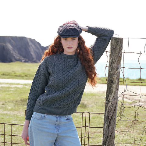 Women's Fisherman Lightweight Crew Neck Heritage Sweater in Lichen, featuring traditional Irish cable knit craftsmanship and heritage-inspired stitching, styled outdoors along the dramatic Irish coastline with rugged cliffs in the background.
