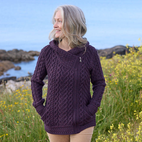 Woman wearing a purple knitted Aran sweater cardigan in a natural setting with rocks and water.