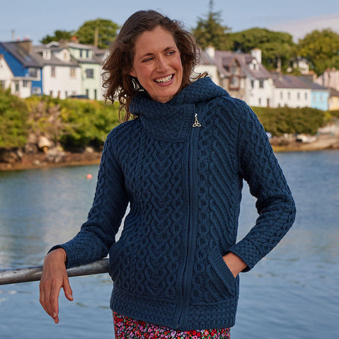 Woman wearing a blue knitted Aran hoodie by a waterfront with houses in the background