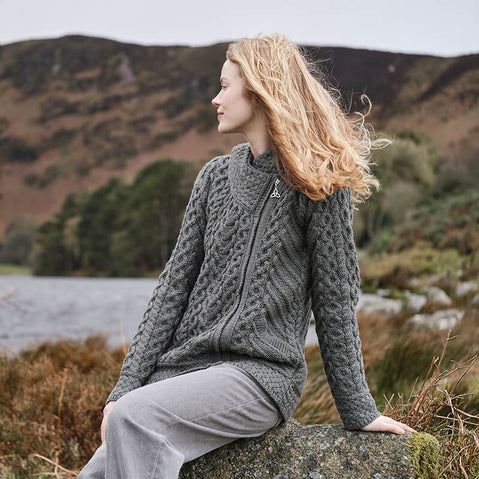A woman standing outdoors wearing a green zippered hooded cardigan with an Aran stitch pattern.