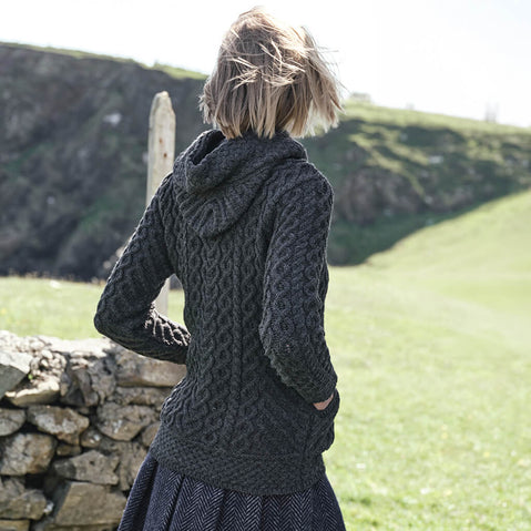Back view of the Women's Signature Aran Celtic Zipper Hooded Cardigan in Charcoal, showcasing intricate traditional Irish cable knit patterns and a textured hood, styled beside a rustic stone wall with the rolling Irish countryside in the background.