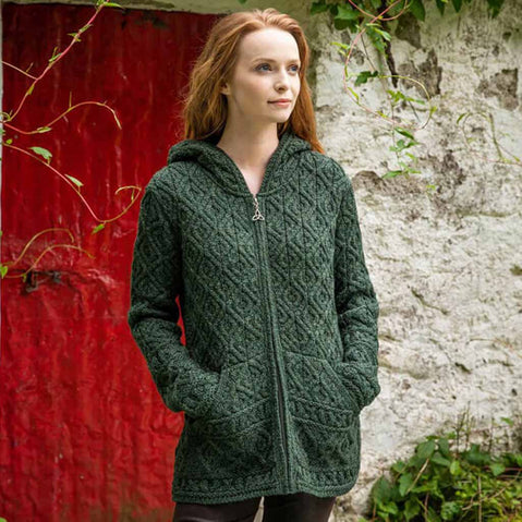 Woman wearing an army green Women's Signature Aran Zipper Hoodie standing against a red wall and stone wall.