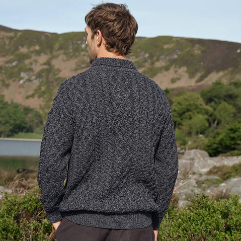Man wearing a dark gray Aran sweater standing in a natural landscape with hills and trees.