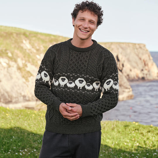 Image featuring the Traditional Sheep Aran Sweater in army green, crafted with authentic Irish Aran cable knit patterns and a classic sheep design inspired by Ireland’s farming heritage, set against a dramatic coastal landscape.