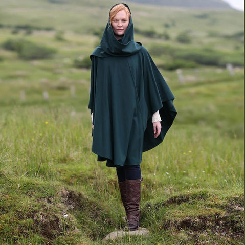 A woman stands in a grassy landscape wearing a dark green cape and brown knee-high boots. She has light hair and is looking directly at the camera, with hills in the background.