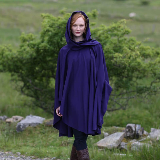 A woman stands outdoors wearing a flowing purple hooded cape. The background features greenery and rocks, suggesting a natural setting. Her expression is calm as she poses in front of the landscape.