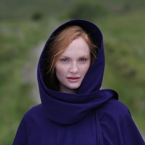 A woman with long red hair is wearing a purple hooded cape. She stands outdoors, with green grass and hilly terrain in the background. The expression on her face is calm and serene.