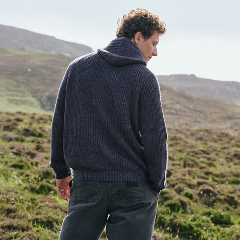 Image featuring the Men's Ribbed Hooded Sweater in indigo, crafted with a traditional ribbed knit design and comfortable hood, showcasing timeless Irish-inspired knitwear from a rear view against a scenic mountain backdrop.