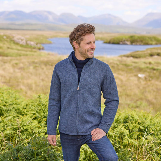 A man standing outdoors wearing a blue Donegal herringbone zipper jacket with long sleeves, paired with jeans.